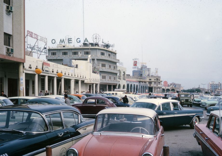 Bustling 1950s city street scene with classic cars parked along the urban road.
